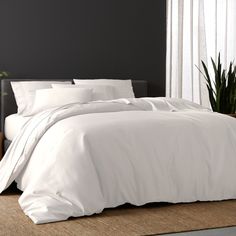 a bed with white sheets and pillows in a room next to a plant on the floor