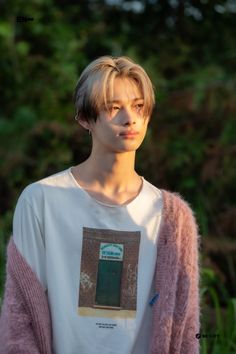 a young man with blonde hair wearing a white shirt and pink cardigan sweater standing in front of trees