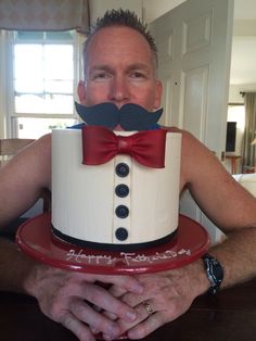 a man holding a cake with a fake mustache on it's face and wearing a red bow tie