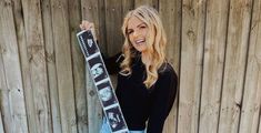 a woman holding up a snowboard in front of a wooden fence with her hand on the board