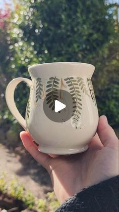 a hand holding a white coffee cup with green leaves on the outside and inside, in front of some bushes