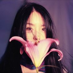 a woman with long black hair holding a flower in front of her face and looking at the camera