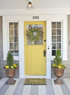 the front door is painted yellow and has two potted plants next to it on the porch