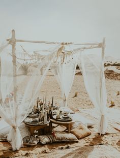 a table set up on the beach for an outdoor dinner party with white drapes