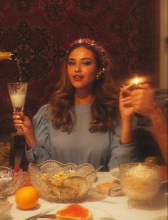 a woman sitting at a table holding a lit candle in front of her face with other food and drinks on the table