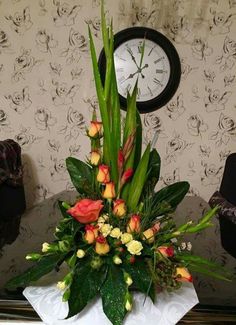 an arrangement of flowers and greenery in front of a clock