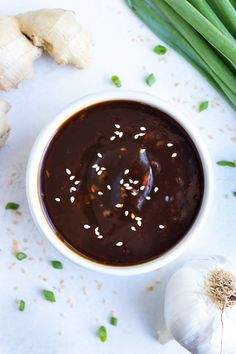 a white bowl filled with brown sauce next to green onions and garlic on a table