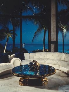 a living room with white couches and palm trees in front of the ocean at night