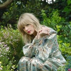 a woman sitting on the ground in front of some bushes and flowers with her hands behind her head