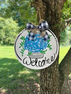 a welcome sign hanging from the side of a tree with blue flowers in a mason jar