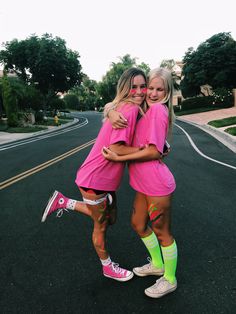 two women in pink shirts and green socks hugging each other on the street with trees behind them