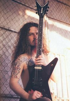 a man with long hair and tattoos holding a guitar in front of a chain link fence