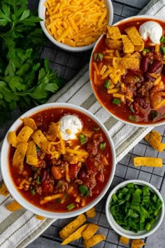 three bowls filled with chili and cheese next to some green beans, sour cream and tortilla chips