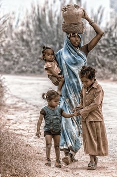 a woman carrying two small children on her back while walking down a dirt road with trees in the background