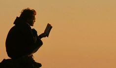 a man sitting on top of a rock reading a book in the sky at sunset