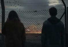 two people standing next to each other in front of a fence with birds flying overhead