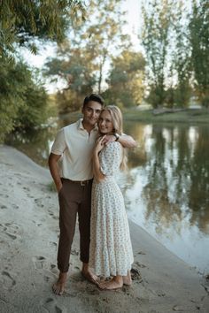 a man and woman standing next to each other on the shore of a lake with trees in the background