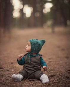 a little boy sitting in the woods wearing a green knitted hat and overalls