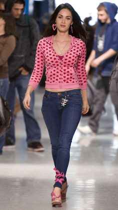 a woman walking down a runway with lots of people in the background wearing jeans and pink sweaters