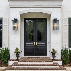 the front entrance to a white brick house with black double doors and two planters