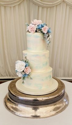 a three tiered wedding cake with pink and blue flowers on the top is sitting on a silver platter