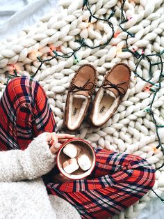 a woman holding a cup of hot chocolate on top of a blanket next to shoes