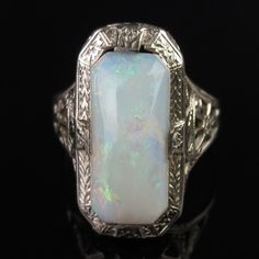 A natural white opal tops this ornate 14k white gold ring by famed jewelers, Ostby & Barton. A carved Art Deco design frames the 13mm x 6mm x 3.8mm cabochon. The white opal has a pinkish undertone with subtle flashes of greens and reds. A pierced filigree design with scrollwork and floral elements decorates the tapered shoulders. The inside of the ring is signed, "OB" (hallmark attributed to Ostby & Barton) followed by "14k" (white gold). The ring measures a petite size 4 (a pinky size for some) Elegant Hallmarked Opal Ring, Classic Opal Moonstone Anniversary Ring, Classic Moonstone Opal Ring For Anniversary, White Platinum Jewelry With Intricate Design, Heirloom White Opal Ring, Classic White Cabochon Opal Ring, Vintage White Opal Ring For Formal Occasions, Classic Opal Anniversary Ring, Heirloom Style Cabochon Opal Ring For Formal Occasions