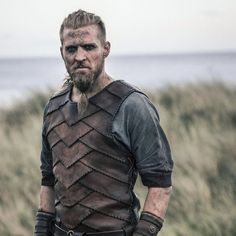 a man with a beard and leather armor standing in front of some grass near the ocean