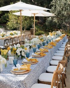 an outdoor dining table set up with blue and white linens, yellow napkins, gold place settings, and umbrellas