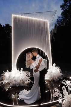 a bride and groom standing in front of a waterfall at night with their arms around each other