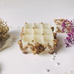 a white and gold jewelry box sitting on top of a table next to purple flowers