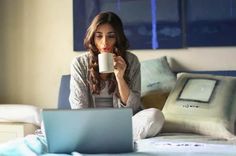 a woman sitting on her bed holding a coffee cup and looking at the laptop screen