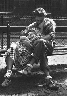 a man and woman sitting on a bench in front of a fence, one holding the other's head