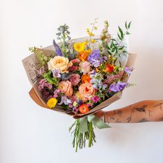 A tattooed arm holds a bouquet wrapped in floral newspaper and brown kraft paper in front of a white backdrop. It's filled with pink, orange, yellow, and purple flowers, including piccolini daisies, roses, veronica, sweet pea, clematis, delphinium, oncidium orchid, and lisianthus. Wildflower Bridal Bouquets, Wildflower Wedding Bouquet, Colorful Wedding Bouquet, Summer Wedding Bouquets, Boquette Flowers, Spring Wedding Flowers, Summer Bouquet, Colorful Bouquet, Wildflower Bouquet