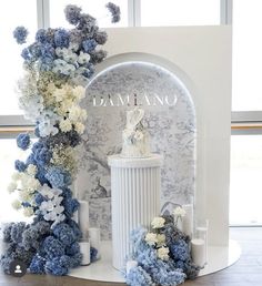 a white and blue wedding cake on top of a pedestal with flowers around the base