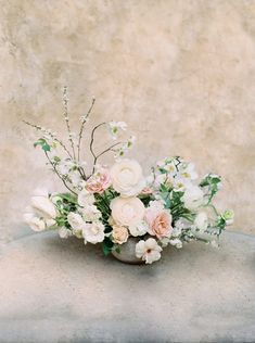 an arrangement of white and pink flowers in a vase on a stone table with a wall behind it