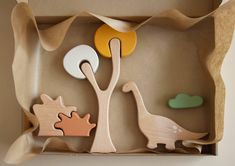 an open cardboard box with wooden toys in the shape of dinosaurs and trees on it