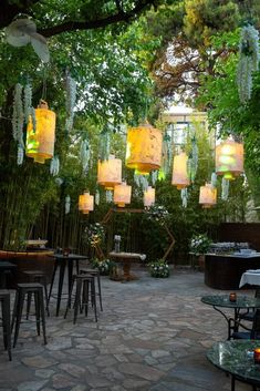 an outdoor dining area with hanging lanterns and tables in the middle of it, surrounded by greenery