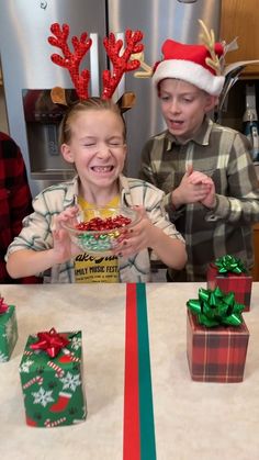 two young boys wearing reindeer antlers and eating christmas treats at a table with presents