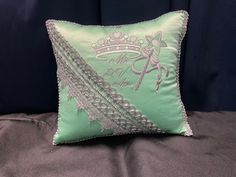 a green pillow with silver trim on it and a tiara in the middle is sitting on a bed