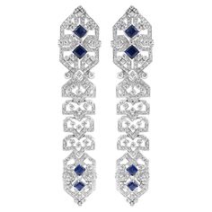 Stunning 18 karat white gold earrings adorned with diamonds and blue sapphires. The intricate design showcases a symmetrical pattern with princess-cut blue sapphires set in the centre, surrounded by sparkling diamonds. The geometric detailing adds a unique and luxurious touch, making these earrings a perfect statement piece for any elegant occasion. Diamond Total Weight: 6.4 carats All Mozafarian jewellery comes with a valuation certificate. Luxury White Gold Diamond Earrings With Intricate Design, Luxury White Gold Sapphire Diamond Earrings, Evening Sapphire Earrings With Diamond Accents, Evening Earrings With Sapphire And Diamond Accents, Sapphire Earrings With Diamond Accents For Evening, Luxury Platinum Blue Earrings, Luxury Blue Platinum Earrings, Formal White Gold Sapphire Diamond Earrings, Elegant Blue Platinum Earrings