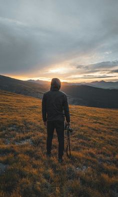 a man standing on top of a grass covered field next to a mountain under a cloudy sky