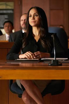 a woman sitting at a desk with other people in the room behind her looking off into the distance