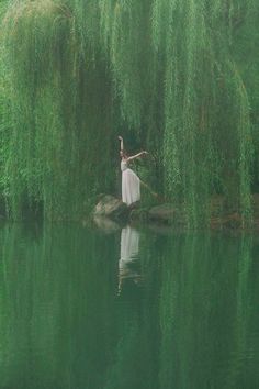 a woman in a white dress is standing on a rock by the water with willow branches