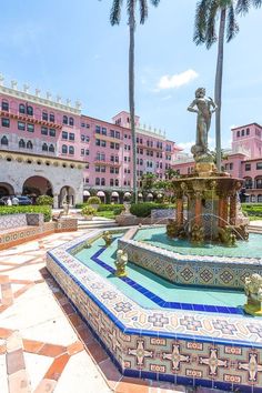 a fountain in the middle of a courtyard with palm trees and pink buildings behind it