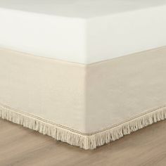 a white bed skirt with fringe trim on the bottom and bottom edge, laying on top of a wooden floor