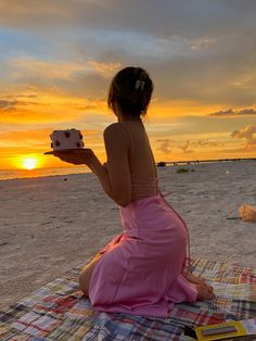 a woman in a pink dress sitting on the beach holding a piece of cake at sunset