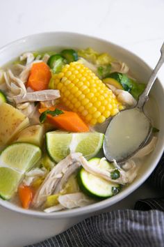 a white bowl filled with chicken, vegetables and corn on the cob next to a spoon