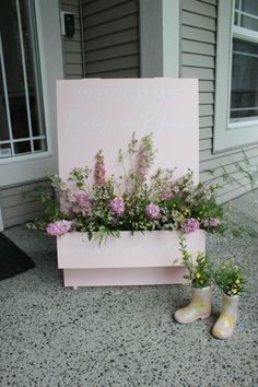 a pink welcome sign with flowers and rain boots sitting on the ground next to it