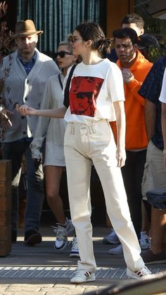 a woman in white pants and a red shirt is standing on the sidewalk with other people behind her